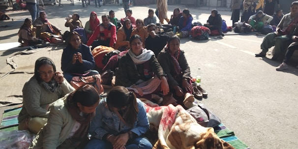 A group of people sitting closely together, many wrapped in colorful blankets and wearing warm clothing. They appear to be gathered on a street or open area, with bags and personal belongings placed around them. The sunlight casts strong shadows, indicating a sunny day.