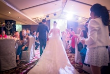A bride and groom walk down an aisle in a warmly lit function room, surrounded by guests seated at tables. Many guests are smiling and capturing the moment with their phones. A woman with a microphone stands to the side, possibly making an announcement or speech. The atmosphere is festive and filled with celebratory energy.