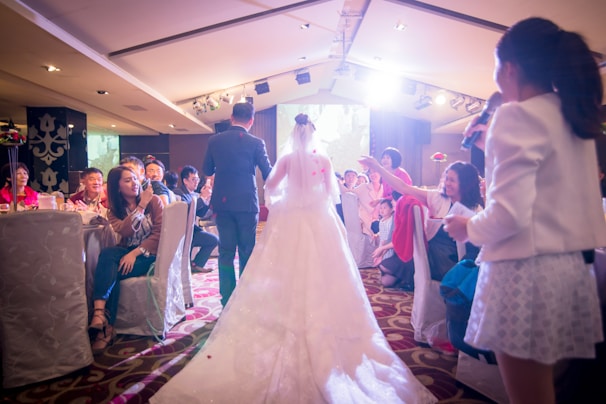 A bride and groom walk down an aisle in a warmly lit function room, surrounded by guests seated at tables. Many guests are smiling and capturing the moment with their phones. A woman with a microphone stands to the side, possibly making an announcement or speech. The atmosphere is festive and filled with celebratory energy.