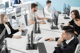 A modern office setting with five people seated at white desks, each working on computers. The desks are arranged in a U-shape, and everyone appears focused on their screens or engaged with their keyboards. The background features a light blue wall, providing a calm and professional atmosphere.