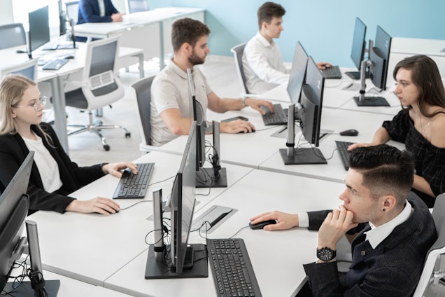 A modern office setting with five people seated at white desks, each working on computers. The desks are arranged in a U-shape, and everyone appears focused on their screens or engaged with their keyboards. The background features a light blue wall, providing a calm and professional atmosphere.