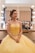 A woman dressed in an elegant yellow gown sits in a brightly lit makeup room. Her hair is styled with colorful flowers, and she looks content. A makeup station with various cosmetics is visible in the background.