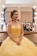 A woman dressed in an elegant yellow gown sits in a brightly lit makeup room. Her hair is styled with colorful flowers, and she looks content. A makeup station with various cosmetics is visible in the background.