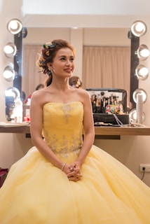 A woman dressed in an elegant yellow gown sits in a brightly lit makeup room. Her hair is styled with colorful flowers, and she looks content. A makeup station with various cosmetics is visible in the background.