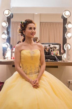 A woman dressed in an elegant yellow gown sits in a brightly lit makeup room. Her hair is styled with colorful flowers, and she looks content. A makeup station with various cosmetics is visible in the background.