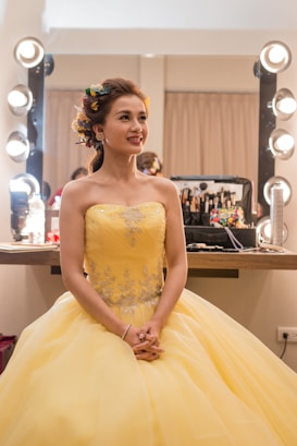 A woman dressed in an elegant yellow gown sits in a brightly lit makeup room. Her hair is styled with colorful flowers, and she looks content. A makeup station with various cosmetics is visible in the background.