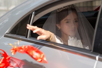 A bride dressed in a white wedding gown and veil is seated in the back seat of a car, reaching out with her hand through the window. She wears a tiara and gold jewelry. A decorative red ribbon adorns the outside of the car.