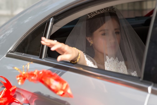 A bride dressed in a white wedding gown and veil is seated in the back seat of a car, reaching out with her hand through the window. She wears a tiara and gold jewelry. A decorative red ribbon adorns the outside of the car.