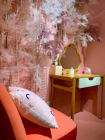 A cozy makeup corner with bright pink walls, white shelves filled with mabê beauty products, and a glowing vanity mirror.