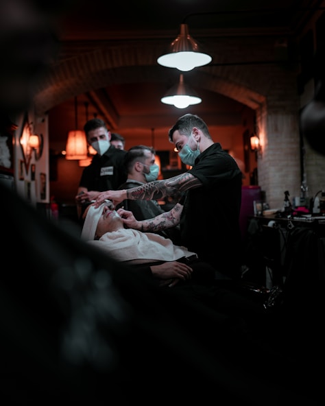 Barbers are wearing masks and attending to clients inside a warmly lit barbershop. One barber is focused on giving a client a shave, while another is visible in the background attending to someone else. The interior is decorated with vintage-style lamps and exposed brick walls, creating a cozy atmosphere.