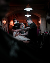 Barbers are wearing masks and attending to clients inside a warmly lit barbershop. One barber is focused on giving a client a shave, while another is visible in the background attending to someone else. The interior is decorated with vintage-style lamps and exposed brick walls, creating a cozy atmosphere.