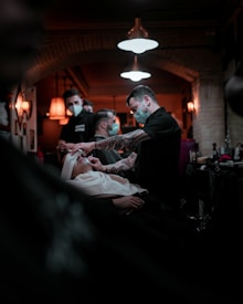 Barbers are wearing masks and attending to clients inside a warmly lit barbershop. One barber is focused on giving a client a shave, while another is visible in the background attending to someone else. The interior is decorated with vintage-style lamps and exposed brick walls, creating a cozy atmosphere.
