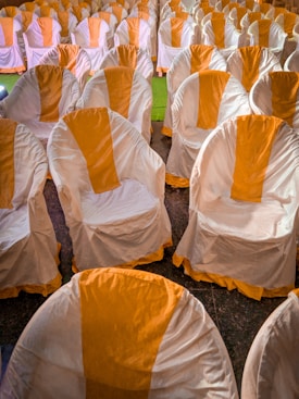 A large number of chairs covered with white cloth and orange accents are arranged in neat rows in an indoor setting that appears to be for an event or ceremony.