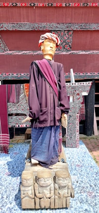 A wooden sculpture of a person wearing traditional clothing with a headdress. The figure stands on a carved wooden base featuring smaller carved figures. The background includes intricate patterns and woven textiles.