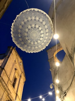 A traditional lace doily is suspended above a street, with its intricate patterns visible against the backdrop of a deep blue evening sky. The scene includes the upper parts of historic buildings, illuminated by soft, hanging string lights that create a warm ambiance.