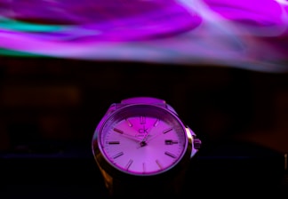 A close-up of a wristwatch with a sleek, modern design. The watch face displays Roman numeral hour markers and three watch hands. The background features swirling patterns of vibrant colors, primarily purple and pink, creating a dynamic and artistic atmosphere.