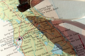 A close-up view of a map featuring the state of Florida, highlighting cities such as Orlando, Tampa, and St. Petersburg. A magnifying glass is partially visible, casting a shadow over the map and emphasizing certain areas.