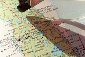 A close-up view of a map featuring the state of Florida, highlighting cities such as Orlando, Tampa, and St. Petersburg. A magnifying glass is partially visible, casting a shadow over the map and emphasizing certain areas.