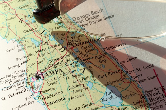 A close-up view of a map featuring the state of Florida, highlighting cities such as Orlando, Tampa, and St. Petersburg. A magnifying glass is partially visible, casting a shadow over the map and emphasizing certain areas.