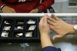 A person is trying on a gold ring at a jewelry counter with a display case containing multiple rings. The person also wears a gold watch, and another individual is visible in the background dressed in a red top.