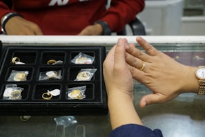 A person is trying on a gold ring at a jewelry counter with a display case containing multiple rings. The person also wears a gold watch, and another individual is visible in the background dressed in a red top.