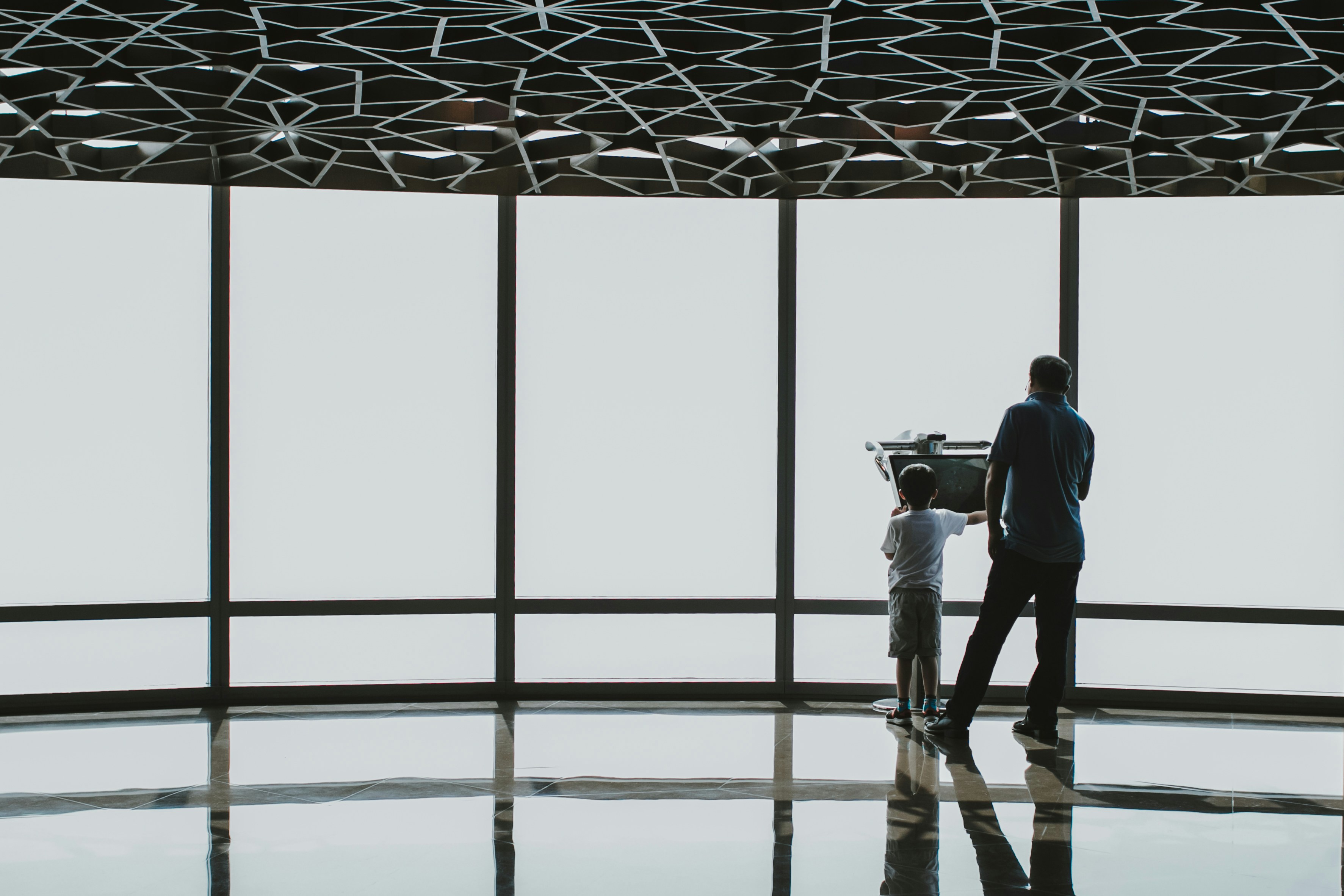a man standing next to a little boy in front of a window, Family using a digital electronic telescope of the Burj Khalifa at the observation deck