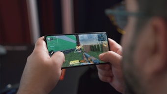 A person is holding a smartphone horizontally and playing a video game. The screen displays a first-person shooter game with a character aiming a gun at targets. The background behind the person is blurred.