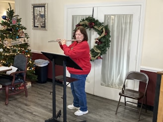 A person wearing a red sweater plays the flute while standing next to a decorated Christmas tree. The setting includes a music stand and folding chair on a wooden floor. The room features holiday decorations, including a large wreath on a door. There is a painting on the wall and another chair with papers on it.