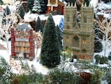 A detailed diorama depicting a snowy village scene during the holiday season. Prominent miniature buildings include a hospital and a cathedral, both adorned with festive decorations such as wreaths. The landscape features snow-covered pine trees and realistic shrubbery, with small human figurines dressed in winter attire engaged in activities around the buildings.