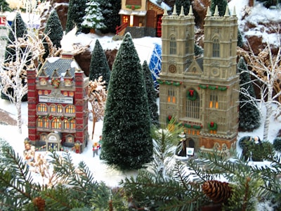 A detailed diorama depicting a snowy village scene during the holiday season. Prominent miniature buildings include a hospital and a cathedral, both adorned with festive decorations such as wreaths. The landscape features snow-covered pine trees and realistic shrubbery, with small human figurines dressed in winter attire engaged in activities around the buildings.