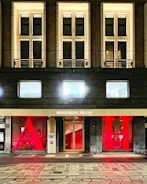 The facade of a hotel building with large, illuminated windows and a modern design. The entrance features a gold sign reading 'Armani Hotel Milano' and is flanked by large red letters 'A' and 'H'. The architecture is sleek with warm lighting emanating from the windows.