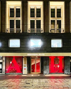 The facade of a hotel building with large, illuminated windows and a modern design. The entrance features a gold sign reading 'Armani Hotel Milano' and is flanked by large red letters 'A' and 'H'. The architecture is sleek with warm lighting emanating from the windows.