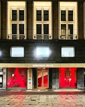 The facade of a hotel building with large, illuminated windows and a modern design. The entrance features a gold sign reading 'Armani Hotel Milano' and is flanked by large red letters 'A' and 'H'. The architecture is sleek with warm lighting emanating from the windows.