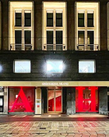 The facade of a hotel building with large, illuminated windows and a modern design. The entrance features a gold sign reading 'Armani Hotel Milano' and is flanked by large red letters 'A' and 'H'. The architecture is sleek with warm lighting emanating from the windows.