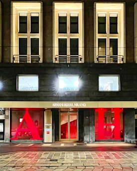 The facade of a hotel building with large, illuminated windows and a modern design. The entrance features a gold sign reading 'Armani Hotel Milano' and is flanked by large red letters 'A' and 'H'. The architecture is sleek with warm lighting emanating from the windows.