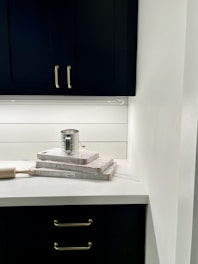 A kitchen countertop features a neatly arranged stack of three wooden cutting boards with a metal can placed on top. A rolling pin rests beside them. The background showcases modern, dark cabinetry with metallic handles and a white wall with horizontal paneling.