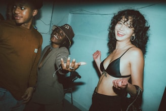 Three people are in a room with a blue-toned wall, engaging in a lively and fun interaction. The woman on the right is smiling and reaching out, wearing a black top, while the man on the left is looking away with a slight smile. The person in the middle is wearing a hat and smiling broadly with an extended arm.