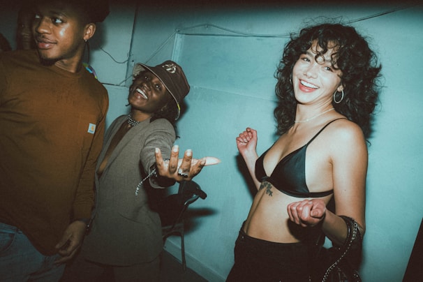 Three people are in a room with a blue-toned wall, engaging in a lively and fun interaction. The woman on the right is smiling and reaching out, wearing a black top, while the man on the left is looking away with a slight smile. The person in the middle is wearing a hat and smiling broadly with an extended arm.