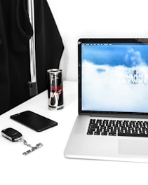 A workspace setup featuring a laptop displaying a cloudy sky on its screen, a can of Red Bull, a smartphone, and a set of keys with a car key fob on a white desk. A black coat or garment is hanging on a metal rack in the background.