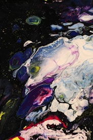 A vibrant abstract painting with swirling patterns featuring bold splashes of color. The image showcases a mix of dark purples and blues, accented with vivid red, pink, and yellow tones. Various shapes seem to overlap and blend into one another, creating a dynamic and fluid composition.