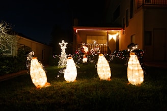 Brightly lit penguin decorations are arranged on a lawn in front of a house, which is also adorned with colorful string lights. A reindeer figure, also illuminated, stands in the background. The setting is nighttime, adding to the festive and cozy atmosphere.
