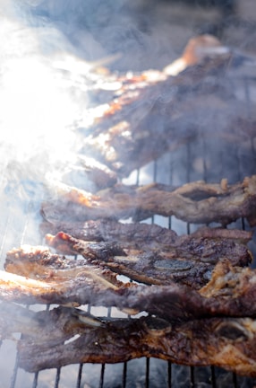 Juicy, brown ribs cooking on an outdoor grill, surrounded by smoke and a burst of sunlight to the left, adding a radiant glow to the scene.