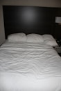 A neatly made bed with three pillows resting against a dark wooden headboard. The bedspread is white and appears slightly wrinkled. Beside the bed, there is a wall-mounted lamp and a portion of a bedside table visible.