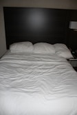 A neatly made bed with three pillows resting against a dark wooden headboard. The bedspread is white and appears slightly wrinkled. Beside the bed, there is a wall-mounted lamp and a portion of a bedside table visible.