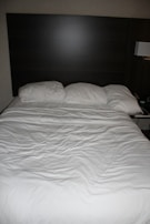 A neatly made bed with three pillows resting against a dark wooden headboard. The bedspread is white and appears slightly wrinkled. Beside the bed, there is a wall-mounted lamp and a portion of a bedside table visible.