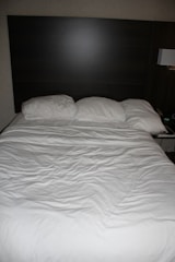 A neatly made bed with three pillows resting against a dark wooden headboard. The bedspread is white and appears slightly wrinkled. Beside the bed, there is a wall-mounted lamp and a portion of a bedside table visible.