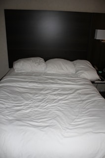 A neatly made bed with three pillows resting against a dark wooden headboard. The bedspread is white and appears slightly wrinkled. Beside the bed, there is a wall-mounted lamp and a portion of a bedside table visible.