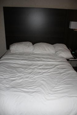 A neatly made bed with three pillows resting against a dark wooden headboard. The bedspread is white and appears slightly wrinkled. Beside the bed, there is a wall-mounted lamp and a portion of a bedside table visible.