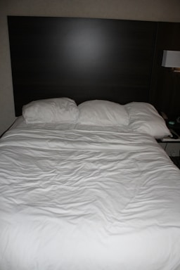 A neatly made bed with three pillows resting against a dark wooden headboard. The bedspread is white and appears slightly wrinkled. Beside the bed, there is a wall-mounted lamp and a portion of a bedside table visible.
