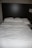 A neatly made bed with three pillows resting against a dark wooden headboard. The bedspread is white and appears slightly wrinkled. Beside the bed, there is a wall-mounted lamp and a portion of a bedside table visible.